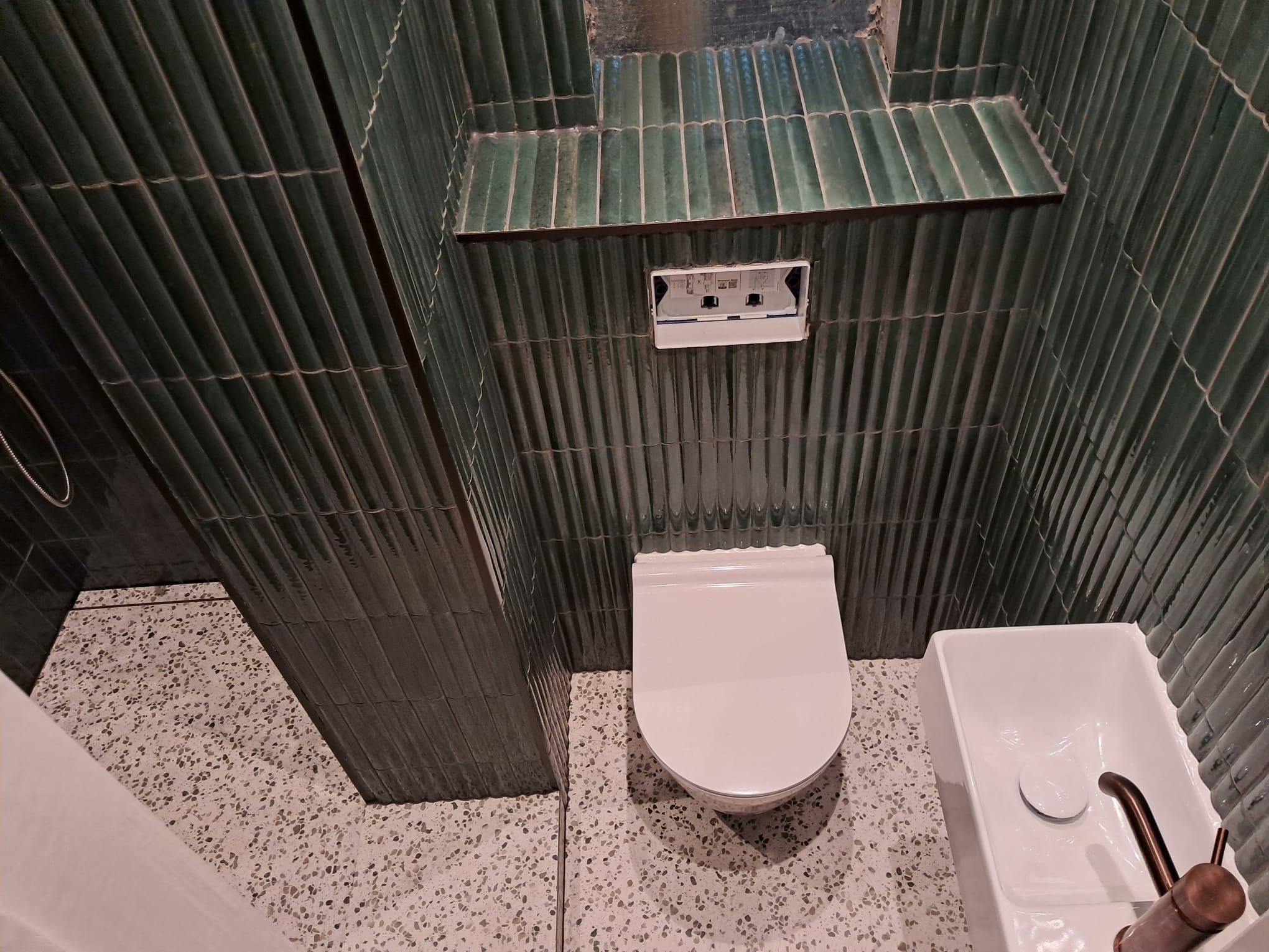 Væghængt toilet og håndvask med grønne vægfliser og lyst terrazzogulv